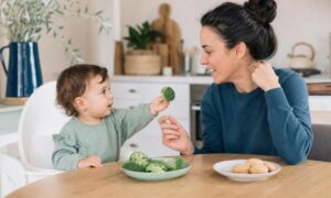 Bambino di diciotto mesi offre un broccolo a un adulto seduto a tavola.