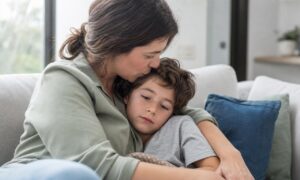 Una madre bacia con premura la fronte del figlio stanco sul divano.